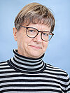 Ein älterer Mensch mit kurzen grauen Haaren, Brille und einem schwarz-weißen gestreiften Rollkragenpullover lächelt leicht. Der Hintergrund ist hellblau.