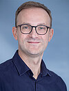 Ein Mann mit kurzen Haaren und einer Brille lächelt freundlich. Er trägt ein dunkelblaues Hemd und hat einen blauen Hintergrund.