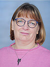Eine Frau mit kurzen, braunen Haaren und einer blauen Brille trägt einen pinken Pullover. Sie lächelt leicht und trägt eine schwarze Halskette mit zwei kleinen Anhängern. Der Hintergrund ist hellblau.