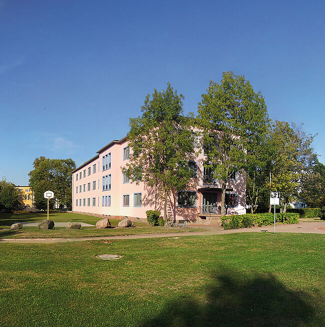 Außenansicht WH 2 Köthen; Foto: Thomas Faust Ein mehrstöckiges Gebäude mit hellrosa Fassade, umgeben von Bäumen und einem gepflegten Rasen. Das Gebäude hat mehrere Fenster und einen zentralen Eingang. Die Umgebung wirkt ruhig und einladend.