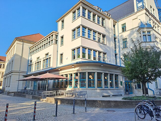 Ein mehrstöckiges Gebäude mit hellen Wänden und vielen Fenstern. Im Erdgeschoss befindet sich ein Café mit einer überdachten Terrasse. Vor dem Gebäude ist ein gepflasterter Bereich mit einigen Bäumen und Fahrradständern. Die Architektur ist modern und ansprechend.