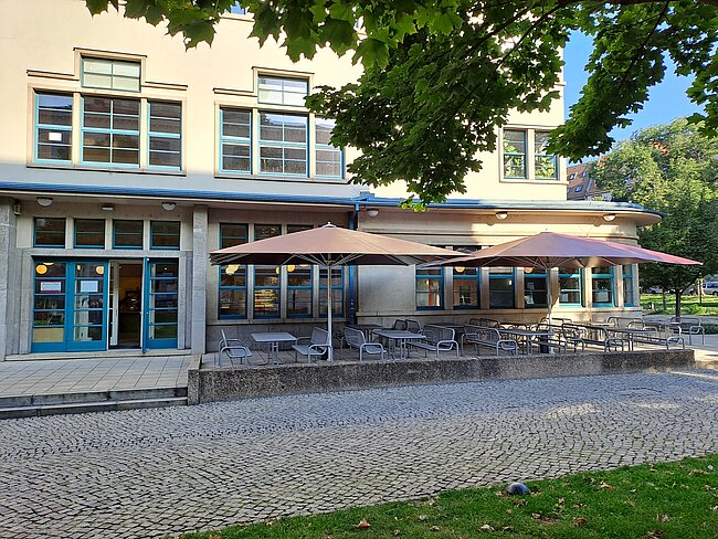 Ein Gebäude mit großen Fenstern und einer Terrasse mit Tischen, Stühlen und Sonnenschirmen. Die Terrasse ist mit Kies gepflastert und von Bäumen umgeben. Im Hintergrund ist ein grasbewachsener Bereich zu sehen.