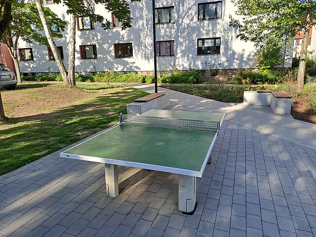 Ein Tischtennistisch steht auf einem gepflasterten Bereich in einem Außenbereich. Im Hintergrund sind Gebäude und Bäume zu sehen. Der Tisch ist bereit für ein Spiel und befindet sich in einem gut gepflegten Umfeld.