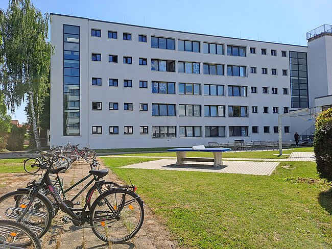 Ein modernes Gebäude mit mehreren Stockwerken und vielen Fenstern. Vor dem Gebäude befindet sich ein Rasen mit einem Tisch und Bänken. Mehrere Fahrräder sind auf dem Gehweg abgestellt. Im Hintergrund ist ein klarer Himmel zu sehen.