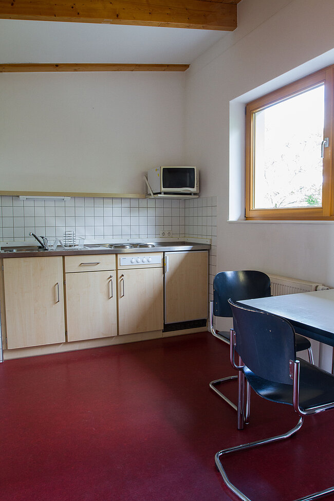 Ein kleiner Küchenbereich mit weißer Arbeitsplatte, zwei Kochplatten, einem Mikrowellenherd und hellen Schränken. Daneben steht ein Tisch mit zwei schwarzen Stühlen. Der Boden ist rot und es gibt ein Fenster mit Holzrahmen.