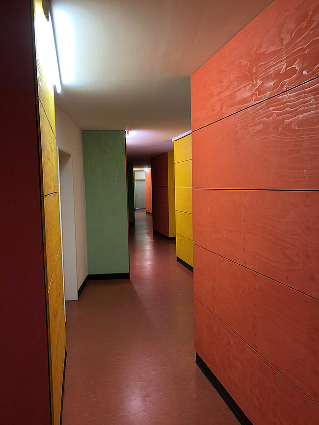 Hausflur WH 2 Köthen; Foto: Thomas Faust Ein langer Flur mit farbigen Wänden in Rot, Gelb und Grün. Die Wände sind mit horizontalen Streifen in verschiedenen Farben gestaltet. Der Boden ist dunkelrot. Am Ende des Flurs ist eine Tür zu sehen. Die Decke hat eine helle Beleuchtung.