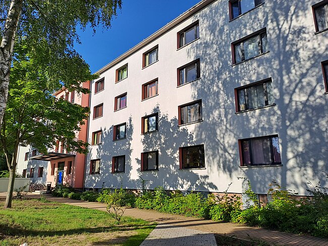 Ein mehrstöckiges Wohngebäude mit weißen Wänden und vielen rechteckigen Fenstern. Vor dem Gebäude befindet sich ein gepflegter Garten mit Bäumen und Sträuchern. Der Himmel ist klar und blau.