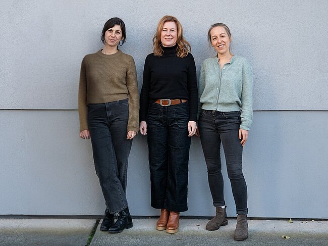 Drei Frauen stehen nebeneinander vor einer grauen Wand. Sie lächeln und tragen lässige, casual Kleidung. Die Frau in der Mitte trägt ein schwarzes Oberteil und eine braune Gürteltasche, die anderen beiden tragen Pullover und Jeans. Sie wirken freundlich und entspannt.