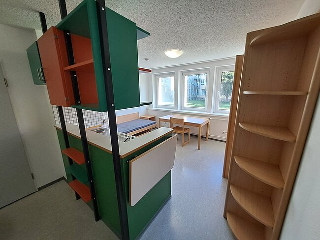 Ein heller Raum mit einem grünen und orangen Regal, einem kleinen Tisch mit Stühlen und einem großen Holzregal. Die Fenster lassen Tageslicht herein, und der Boden ist grau. Der Raum wirkt wie ein Klassenzimmer oder ein Arbeitsbereich.