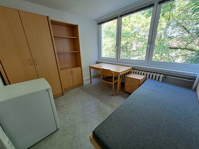 Ein einfaches Zimmer mit einem Bett, einem Schreibtisch, einem Stuhl, einem kleinen Schrank und einem Kühlschrank. Große Fenster bieten viel Tageslicht und Blick auf Bäume. Die Einrichtung ist schlicht und funktional, ideal für ein Studenten- oder Gästezimmer.