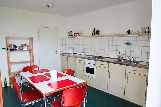Ein helles Küchenzimmer mit weißem Tisch und vier roten Stühlen. Die Küche hat helle Schränke, eine Arbeitsplatte mit Spüle und Herd. An der Wand sind Regale mit Geschirr und Vorräten. Der Boden ist dunkelgrün. Ein Kühlschrank ist in der Nähe des Tisches.