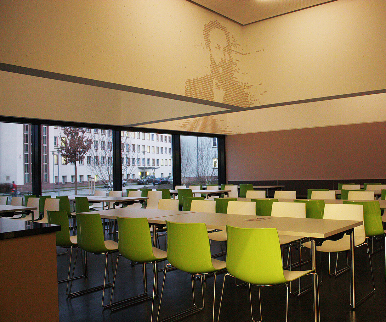 Speisesaal Heidemensa; Foto: Thomas Faust Ein moderner Speisesaal mit mehreren Tischen und grünen Stühlen. Die Wände sind weiß mit einem subtilen Porträt an einer Wand. Große Fenster bieten Blick auf die Straße und Gebäude draußen. Der Raum wirkt hell und einladend.