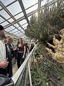 "Menschen besuchen ein Gewächshaus mit verschiedenen Sukkulenten und Kakteen. Die Pflanzen sind in einer natürlichen, felsigen Umgebung angeordnet. Die Besucher stehen hinter einer Absperrung und betrachten die Pflanzen."