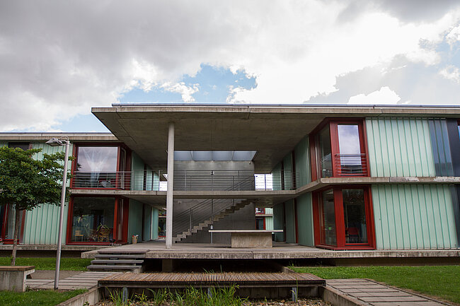 Moderne Architektur mit zwei Gebäuden, die durch eine überdachte Brücke verbunden sind. Die Gebäude haben große Fenster und rote Rahmen. Vor den Gebäuden befindet sich ein grasbewachsener Bereich mit Holzbänken. Der Himmel ist teilweise bewölkt.