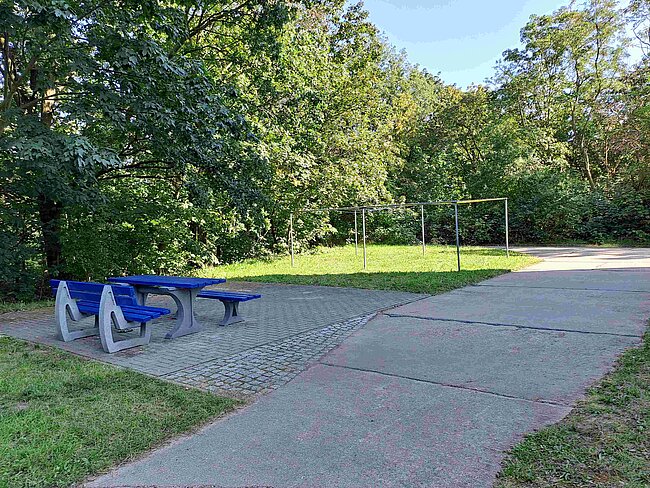 Ein Parkbereich mit zwei blauen Bänken und einem Picknicktisch. Im Hintergrund befindet sich ein kleiner Fußballplatz mit Tor. Der Bereich ist von Bäumen umgeben und hat einen gepflasterten Weg.