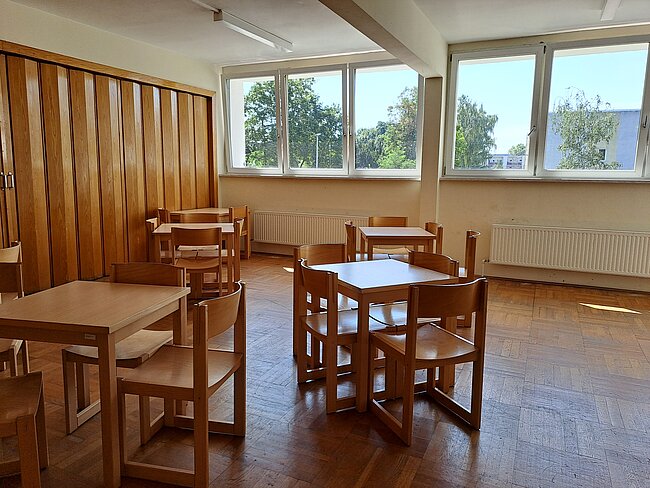 Ein heller Raum mit mehreren Holztischen und Stühlen. Große Fenster lassen Tageslicht herein. An den Wänden befinden sich Heizkörper. Der Boden ist aus Holz. Der Raum wirkt sauber und aufgeräumt, möglicherweise ein Klassenzimmer oder ein Gemeinschaftsraum.