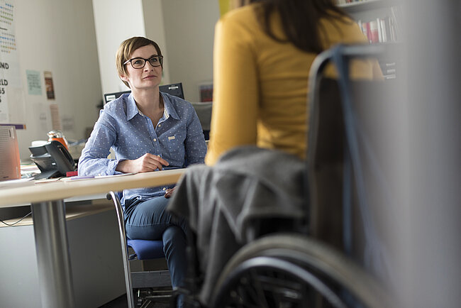 Eine Frau mit Brille sitzt an einem Tisch in einem Büro und schaut nachdenklich. Im Vordergrund ist ein Rollstuhl zu sehen. Eine weitere Person in einem gelben Pullover steht im Hintergrund. Der Raum wirkt wie ein Arbeitsumfeld mit Büromaterial und Regalen.