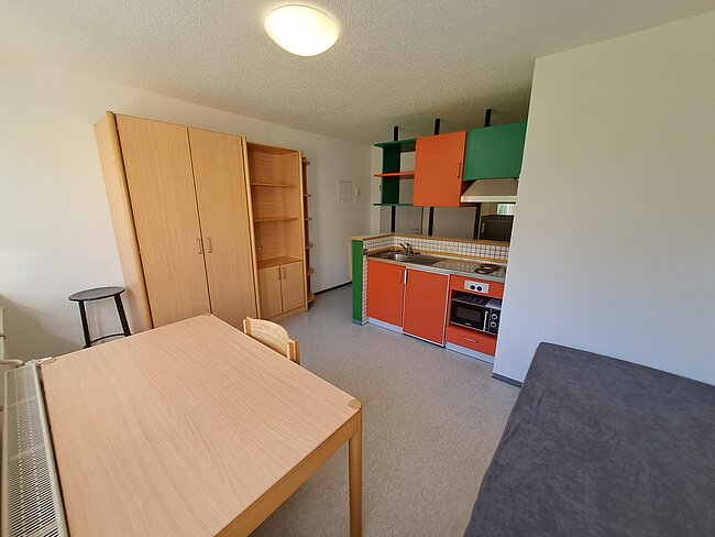 Ein kleiner, hell gestalteter Raum mit Küchenzeile, Esstisch und Bett. Die Küche hat orangen und grünen Schränken mit weißer Arbeitsplatte. Der Esstisch ist aus hellem Holz, und ein graues Bett befindet sich rechts im Raum. Ein großer Schrank steht links neben dem Tisch.