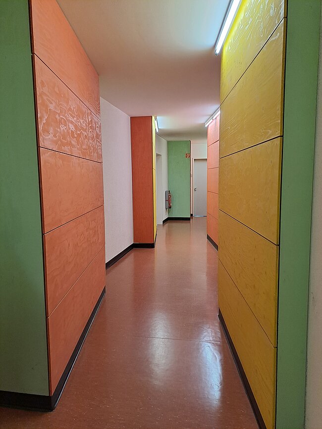 Ein langer Flur mit bunten Wänden in Grün, Orange und Gelb. Der Boden ist rot. Am Ende des Ganges befindet sich eine Tür. Die Decke ist weiß und hat eine Lichtöffnung. Der Flur wirkt sauber und modern.