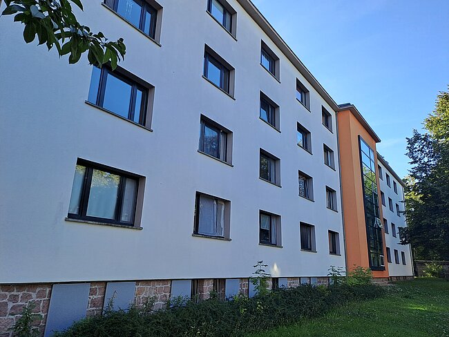 Ein mehrstöckiges Wohngebäude mit weißer Fassade und orangefarbenem Seitenteil. Die Fassade hat zahlreiche rechteckige Fenster. Im Vordergrund befindet sich ein bewachsener Bereich mit Grünflächen. Der Himmel ist klar und blau.