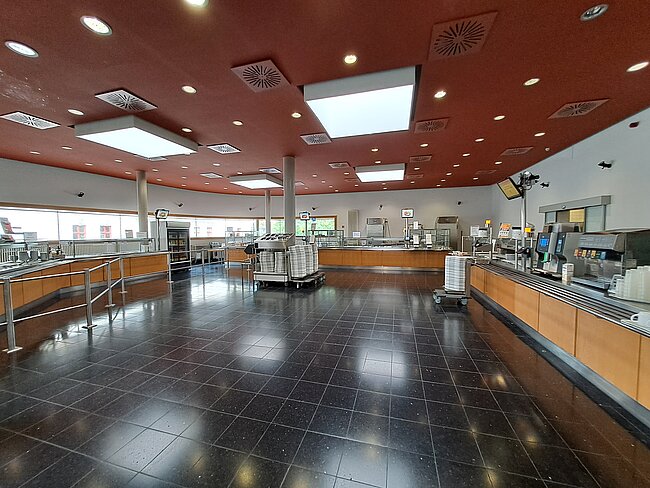 Ausgabebereich Ein großer, sauberer Raum mit glänzendem schwarzen Boden und rotem Deckenbereich. Es gibt mehrere Theken mit verschiedenen Geräten und Ausrüstungen, die auf eine Cafeteria oder Kantine hindeuten. Der Raum ist gut beleuchtet und wirkt einladend, aber derzeit leer.