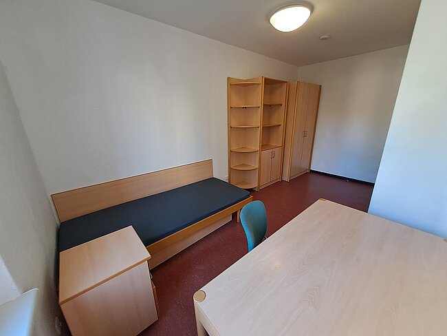 Ein einfaches, spartanisch eingerichtetes Zimmer mit einem schmalen Bett, einem Schreibtisch, einem Stuhl und einem Regal. Die Wände sind weiß, der Boden mit rotem Teppich ausgelegt. Das Zimmer ist gut beleuchtet und wirkt sauber und ordentlich.