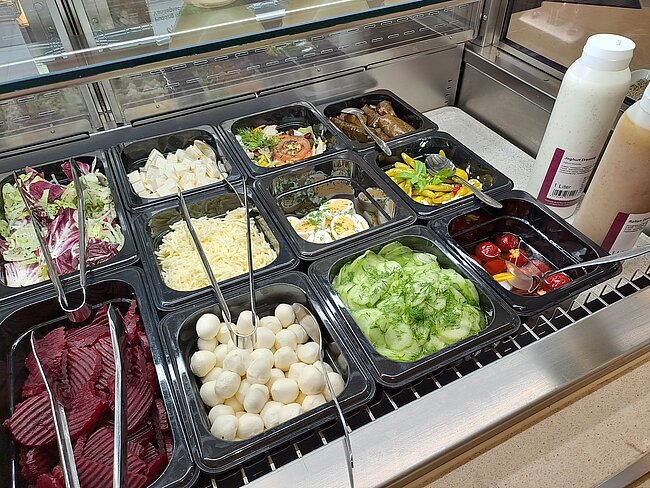 Ein Buffet mit verschiedenen Salatzutaten: Blattsalate, Gurken, Eier, Mais, Bohnen, Fleischstreifen und andere Zutaten in schwarzen Schüsseln. Servierlöffel sind in den Schüsseln platziert.