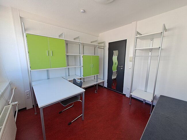 Ein leerer Raum mit grünen Schränken, Metallregalen und einem Tisch. Der Boden ist rot, und es gibt eine Tür mit einem grünen Notausgang-Schild. Der Raum wirkt wie ein Klassenzimmer oder ein Büro, das noch eingerichtet werden muss.