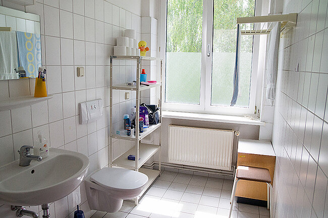 Bad in 5er-WG Wohnheim Magdeburger Straße, Bernburg; Foto: Annegret Höhne Ein modernes Badezimmer mit weiß gefliesten Wänden und Boden. Es enthält ein Waschbecken, eine Toilette, ein Regal mit verschiedenen Toilettenartikeln, einen Heizkörper und ein Fenster mit Vorhängen. Ein Duschvorhang ist ebenfalls sichtbar.