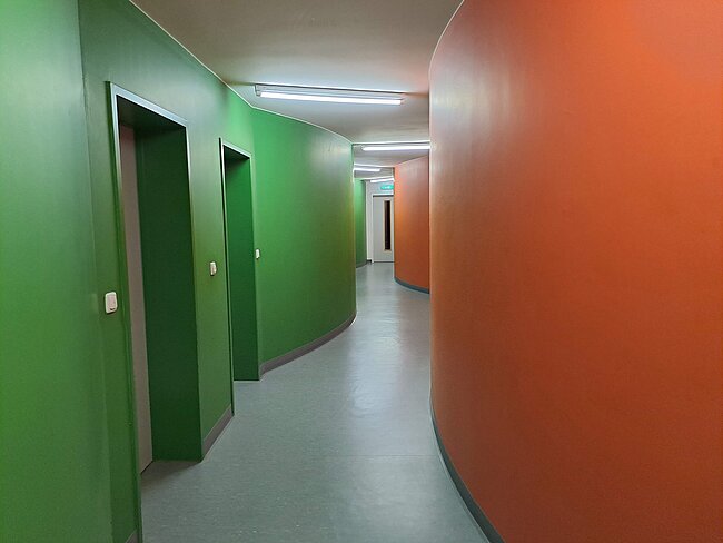 Ein langer Flur mit grünen Wänden auf der linken Seite und einer großen, runden, orangen Wand auf der rechten Seite. Es gibt mehrere Türen auf der grünen Seite und das Ende des Flurs ist in der Ferne sichtbar. Der Boden ist grau und glatt.