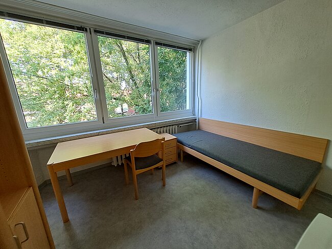 Ein einfaches Zimmer mit einem Bett, einem Schreibtisch und einem Stuhl. Das Zimmer hat große Fenster, durch die man Bäume und Grünflächen sehen kann. Die Wände sind hell und der Boden ist mit grauem Teppichboden bedeckt.