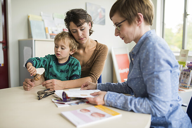 Eine Frau und ein Kind sitzen an einem Tisch und werden von einer anderen Frau beobachtet. Das Kind hält einen Stift und scheint etwas zu malen oder zu schreiben. Vor ihnen liegen bunte Blätter und Bücher. Die Szene wirkt wie eine Lernsituation oder ein kreativer Moment.