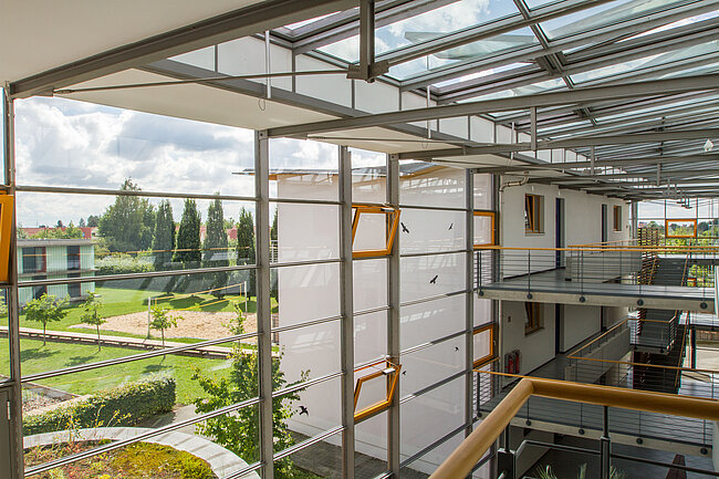 Ein modernes Gebäude mit großen Glasfenstern und einer offenen Treppe. Im Hintergrund ist ein gepflegter Garten mit Bäumen und Wegen zu sehen. Das Design ist hell und luftig mit viel natürlichem Licht.
