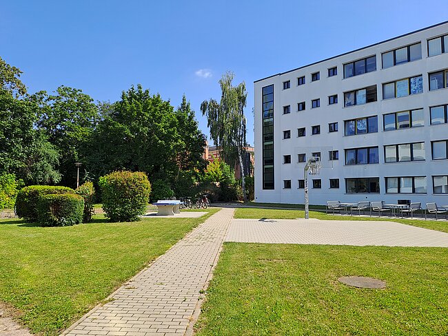 Ein modernes, mehrstöckiges Gebäude mit vielen Fenstern steht auf der rechten Seite. Vor dem Gebäude befindet sich ein gepflegter Rasen mit einem gepflasterten Weg, der zum Eingang führt. Im Hintergrund sind Bäume und Sträucher zu sehen.