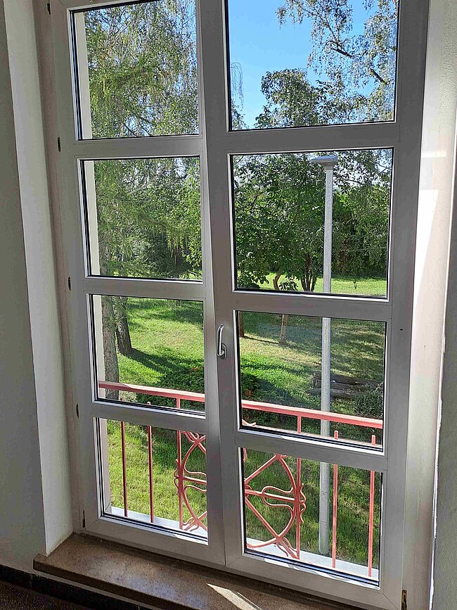 Ein Fenster mit Blick auf einen grünen Garten mit Bäumen und Rasen. Vor dem Fenster befindet sich ein roter Metallgitterzaun. Das Fenster ist in mehrere Scheiben unterteilt und leicht geöffnet.