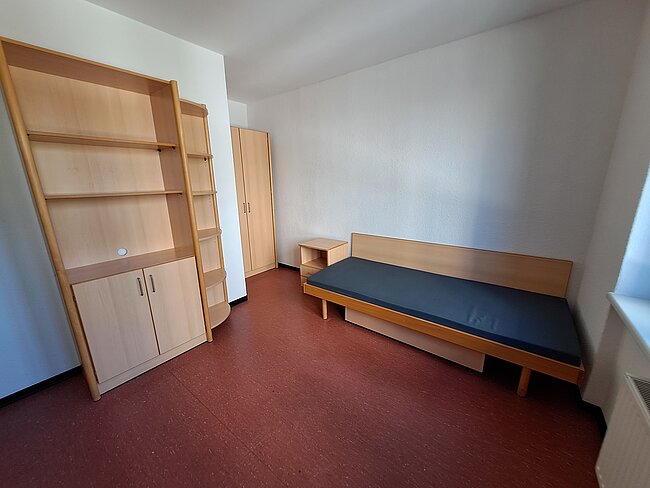 Ein einfaches Zimmer mit einem schmalen Bett, einem kleinen Nachttisch und einem großen Holzregal mit Schubladen und Fächern. Der Boden ist mit rotem Teppich ausgelegt, und die Wände sind weiß. Die Tür führt in einen weiteren Raum oder Flur.