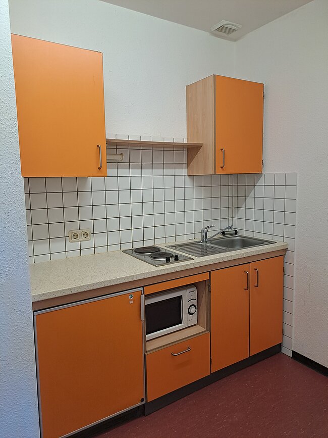 Eine kleine Küche mit orangefarbenen Schränken, weiß gefliesten Wänden und einer Arbeitsplatte. Sie enthält einen Herd mit zwei Kochplatten, eine Spüle und einen eingebauten Mikrowellenofen. Oberhalb der Arbeitsplatte befinden sich zwei Hängeschränke und ein Regal.