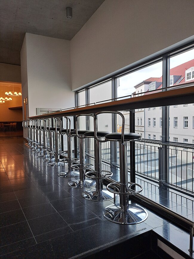 Barhocker und Tresen mit Ausblick auf den Harz Ein langer Tresen mit vielen hohen Hockern steht vor einer großen Glasfront. Durch die Fenster sieht man Gebäude und den Himmel. Der Raum wirkt modern und hell. Der Boden ist dunkel gefliest. Im Hintergrund sind weitere Möbel und ein Kronleuchter zu erkennen.