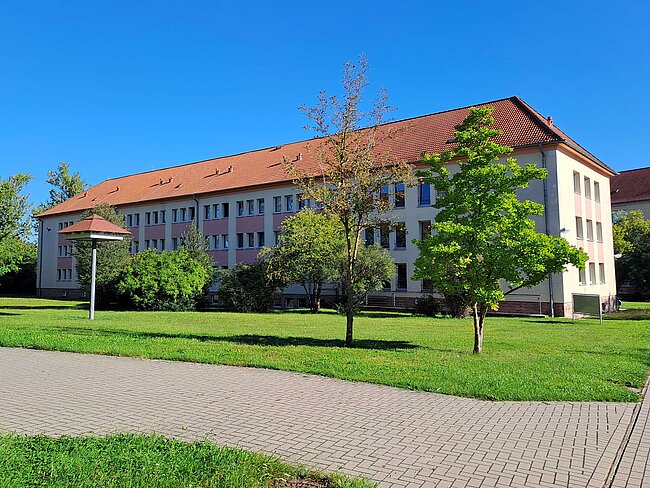Ein großes, weißes Schulgebäude mit rotem Dach und vielen Fenstern. Vor dem Gebäude befindet sich ein gepflegter Rasen mit Bäumen und einem gepflasterten Weg. Der Himmel ist klar und blau.