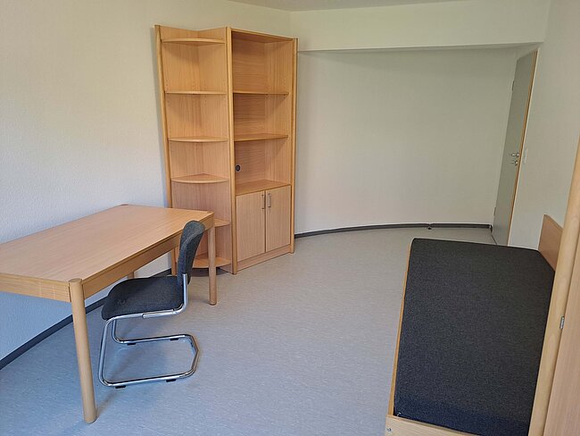 Ein einfaches Zimmer mit einem Schreibtisch, einem Stuhl und einem Bücherregal. An einer Wand befindet sich ein Bett mit einer schwarzen Matratze. Der Boden ist mit grauem Teppichboden bedeckt, und die Wände sind weiß gestrichen.