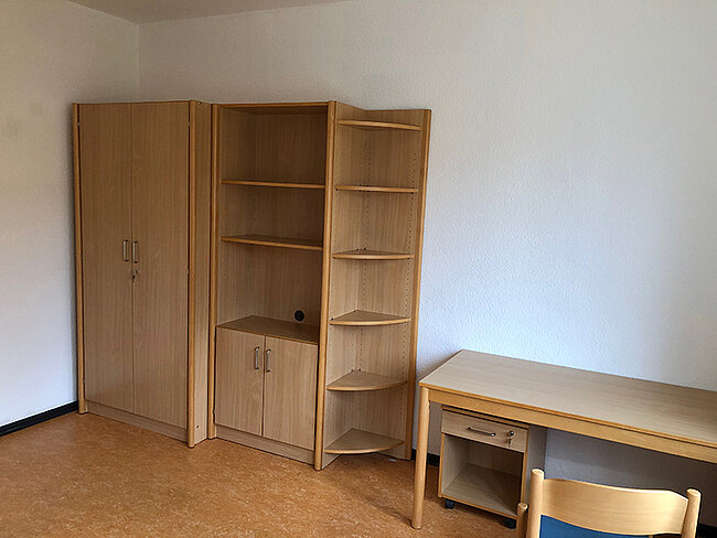 Zimmer WH 2 Köthen; Foto: Thomas Faust Ein einfaches Zimmer mit einem großen Schrank, einem Regal und einem Schreibtisch mit einem Stuhl. Der Schrank hat zwei Türen und mehrere Fächer. Das Regal hat mehrere offene Ablageflächen. Der Schreibtisch ist schlicht und hat eine Schublade. Der Boden ist mit Teppich ausgelegt.