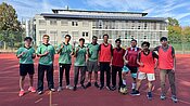 Gruppe von elf Personen auf einem Sportplatz, die fröhlich posieren. Sieben tragen grüne Trikots, vier rote. Im Vordergrund liegt ein Fußball.