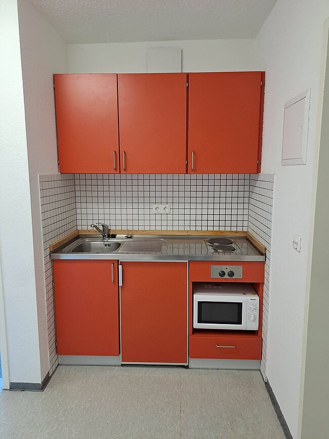 Ein kleiner Küchenbereich mit orangefarbenen Schränken und Arbeitsplatte. Es gibt eine Spüle, einen Herd mit zwei Kochplatten und einen eingebauten Mikrowellenofen. Die Wände sind mit weißen Fliesen hinter dem Arbeitsbereich verkleidet. Der Boden ist hellgrau.