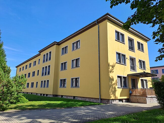 Ein mehrstöckiges, gelbes Gebäude mit vielen Fenstern und einem kleinen Vordach am Eingang. Das Gebäude steht auf einem gepflasterten Weg umgeben von Grünflächen und Bäumen. Der Himmel ist klar und blau.