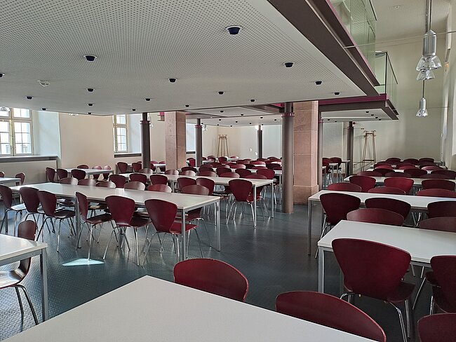 Ein großer, leerer Speisesaal mit vielen Tischen und roten Stühlen. Die Tische sind in Reihen angeordnet und der Raum wirkt sauber und ordentlich. Große Fenster lassen Tageslicht herein und die Decke hat mehrere Lampen.