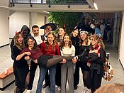Eine Gruppe junger Menschen feiert zusammen in einem Raum. Sie tragen lustige Kostüme und Accessoires, darunter Teufelshörner, Hexenhüte und andere Halloween-Artikel. Die Stimmung wirkt fröhlich und ausgelassen, während sie für ein Gruppenfoto posieren.