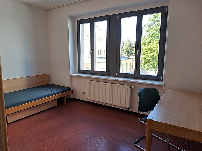 Ein einfaches Zimmer mit einem schmalen Bett, einem Schreibtisch mit Stuhl und einem großen Fenster. Das Fenster hat vier Flügel und bietet einen Blick auf die Umgebung. An der Wand befindet sich ein Heizkörper. Der Boden ist mit einem Teppich bedeckt.