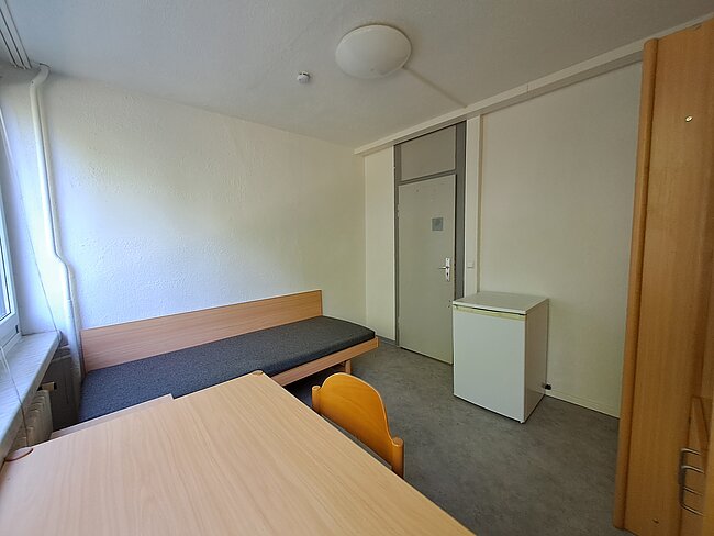 Ein einfaches Zimmer mit einem Tisch und zwei Stühlen, einem schmalen Bett, einem kleinen Kühlschrank und einer Tür. Die Wände sind weiß, und es gibt ein Fenster auf der linken Seite. Der Boden ist mit grauem Teppich bedeckt.