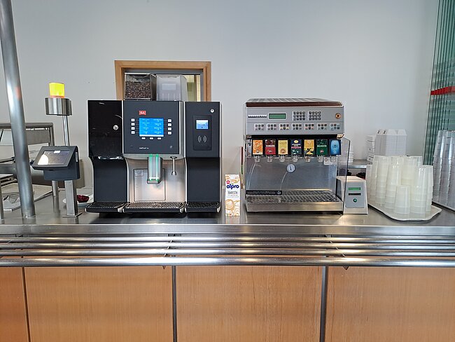 Kaffeeautomat und Getränkeautomat Zwei Kaffeemaschinen stehen nebeneinander auf einer Theke. Die linke Maschine hat einen Bildschirm und Knöpfe, während die rechte Maschine mit verschiedenen Tasten und einem Milchaufschäumer ausgestattet ist. Beide Maschinen sind bereit, Kaffee zuzubereiten.