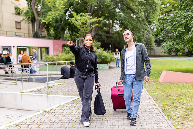 Wohnheimturor/innen helfen beim Ankommen und Orientieren Zwei Personen gehen auf einem Weg in einem Park oder Campusgelände. Eine Frau zeigt nach links und trägt eine schwarze Tasche, während ein Mann einen pinken Koffer hinter sich herzieht. Beide wirken entspannt und unterhalten sich.
